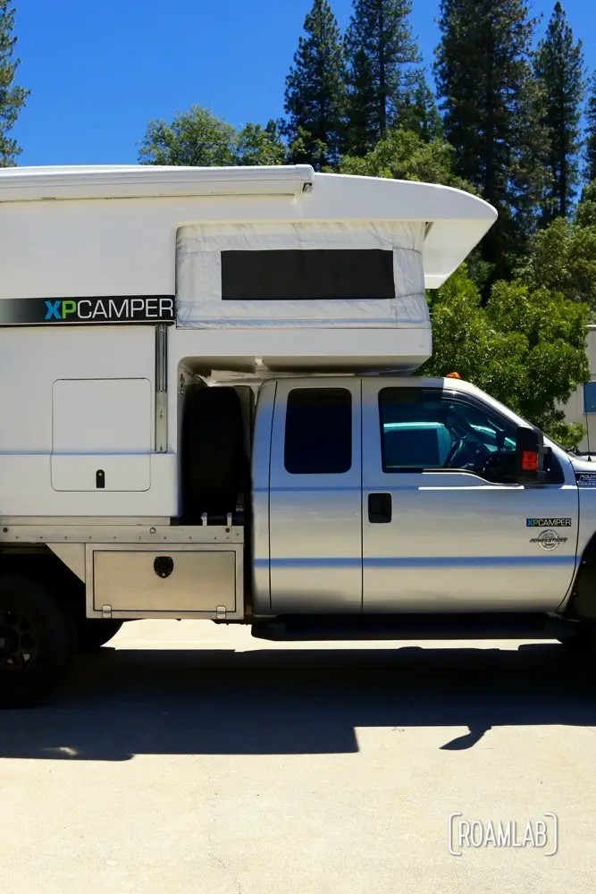 This is a fully expanded XP Camper. The section above the truck cab is fabric but everything else has hard siding. You can see a line along the center of the body where the top section slides over the bottom section when the camper is compressed. The side entrance allows a full width table with picture window at the back but means that this camper is not a slide in. The truck bed is converted to allow a full width camper. See the industrial metal base that is substituted for a truck bed.