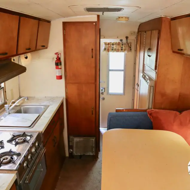 Interior view of a 1970 Avion C11 truck camper featuring a kitchen counter top, dinette table, stove, sink, refrigerator, overhead storage, and exit door.