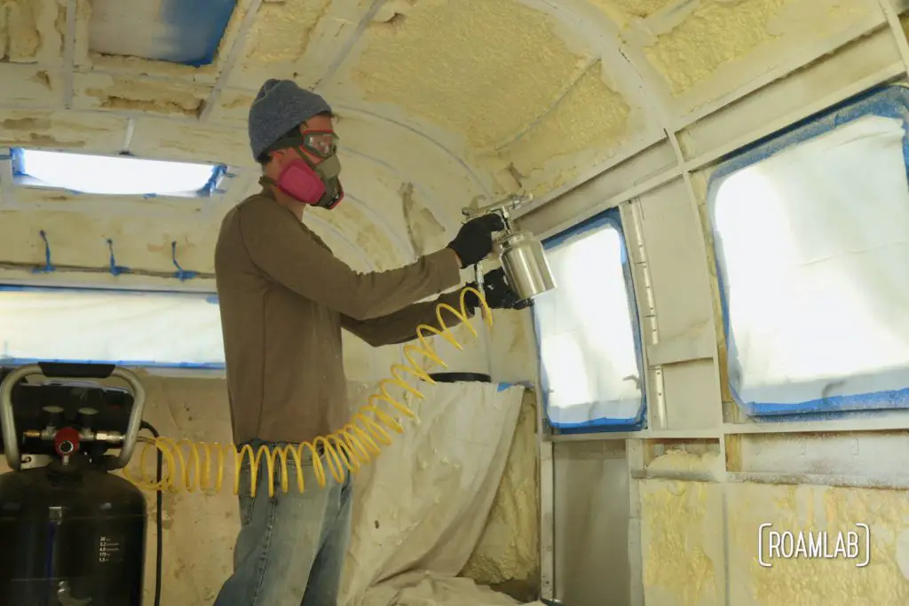 Man applying white paint inside a camper with a spray gun attached to an air compressor.