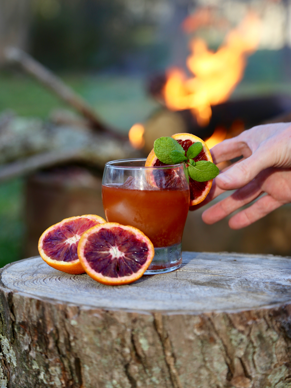 Hand reaching for a glass of blood orange cocktail with a fire in the background
