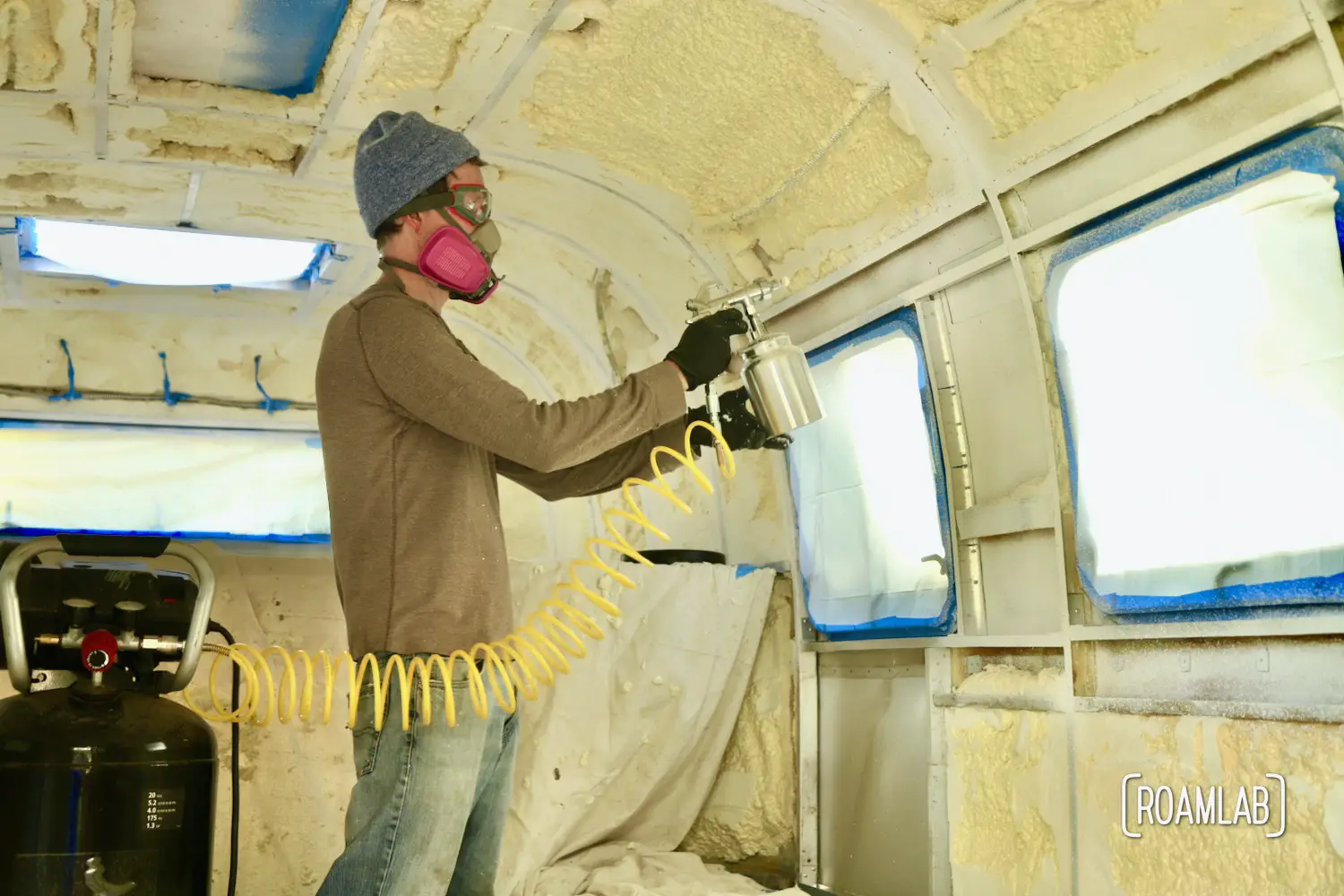 Man applying white paint inside a camper with a spray gun attached to an air compressor.
