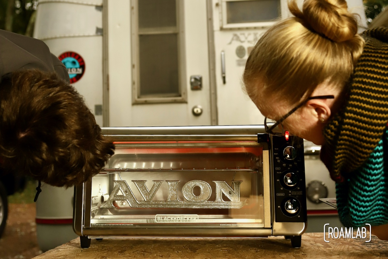 Man and woman watching an Avion emblem with a chrome powder coat cure in a toaster oven.