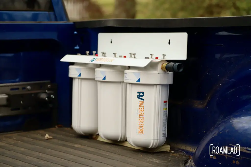 A three container water filter system propped in place on a board of wood inside a truck bed.