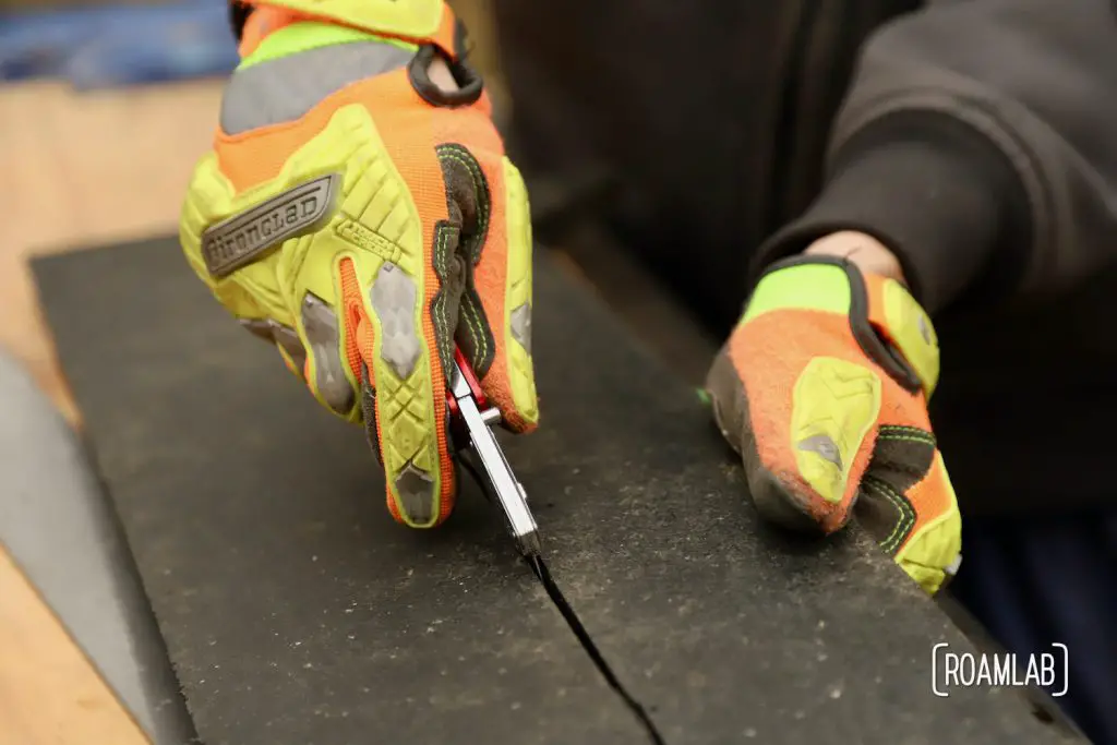 Gloved hands cutting a thick piece of black rubber with a box cutter.