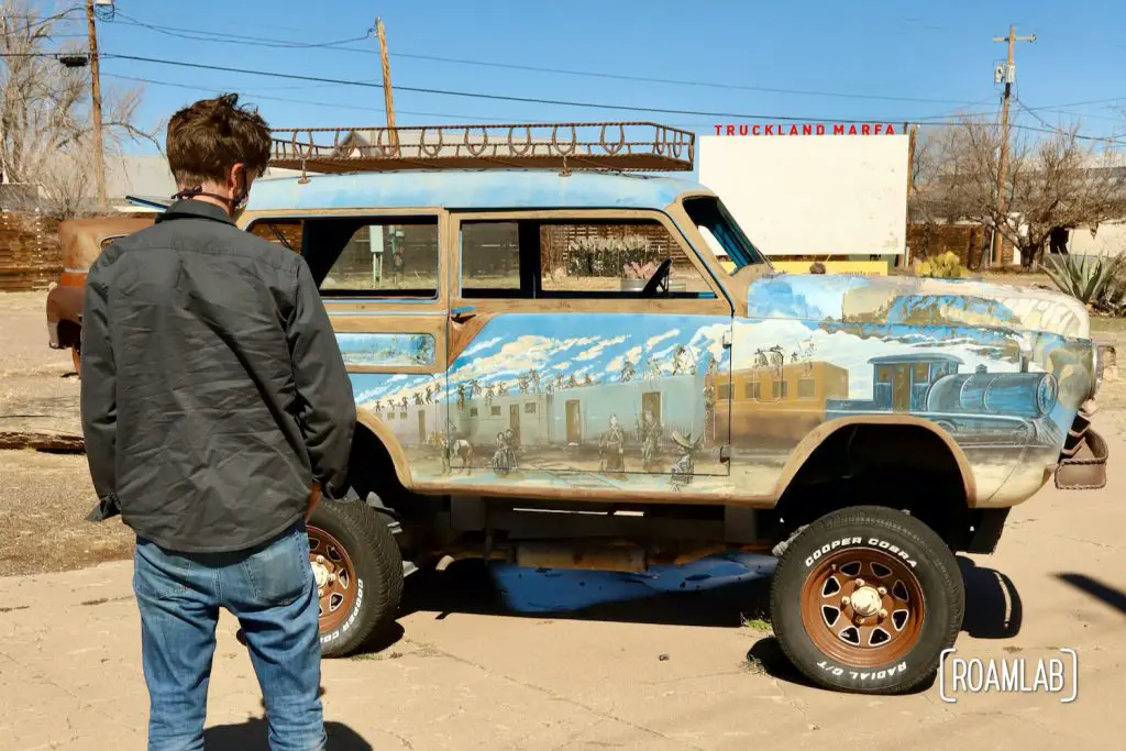 Man standing in front of a mural painted on the side of an old car with a sign for Truckland Marfa in the background.