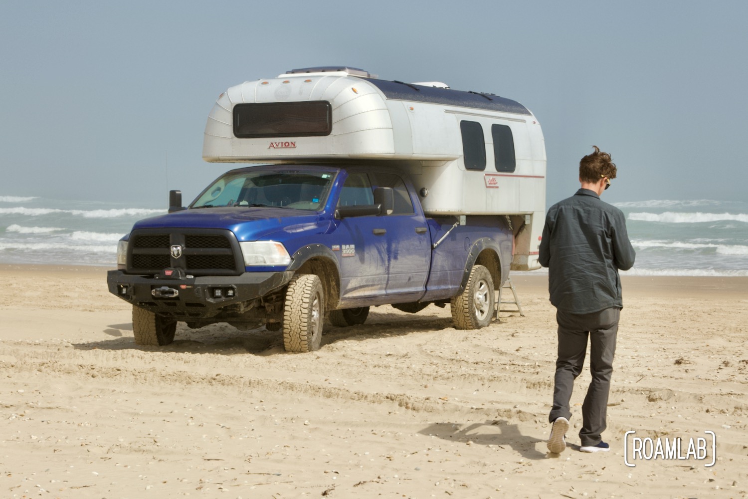 Man walking in front of a 1970 Avion C11 truck camper parked on the beach in South Padre Island, Texas