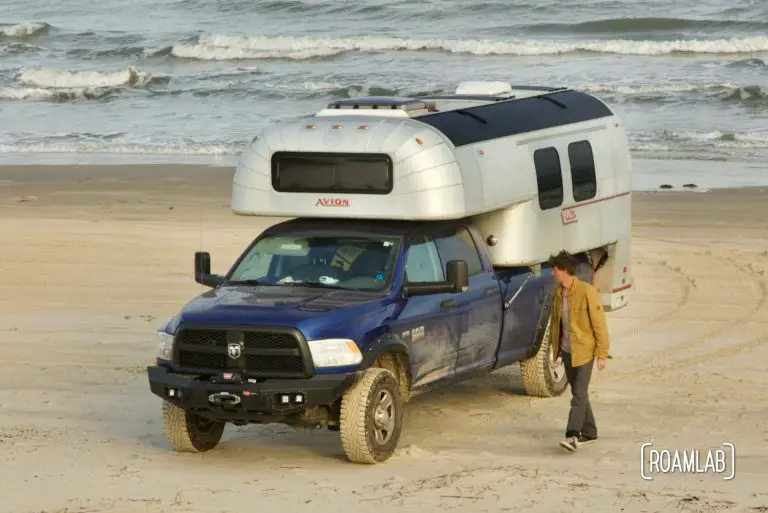 Man walking along the beach, inspecting a 1970 Avion C11 truck camper parked by the water at South Beach Padre Island National Seashore, Texas.