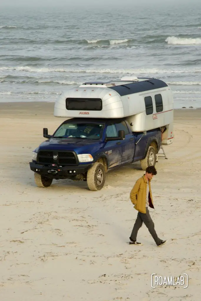 Man walking along the beach past at 1970 Avion C11 truck camper parked by the water at South Beach Padre Island National Seashore, Texas.
