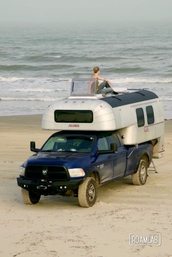 Woman sitting on the roof of a 1970 Avion C11 truck camper parked on the beach at South Beach Padre Island National Seashore, Texas.