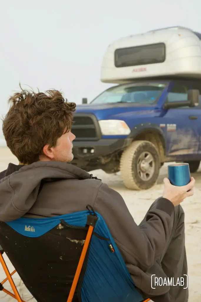 Man sitting in a collapsible camping chair on the beach, holding a blue glass in front of a 1970 Avion C11 truck camper at South Beach Padre Island National Seashore, Texas.