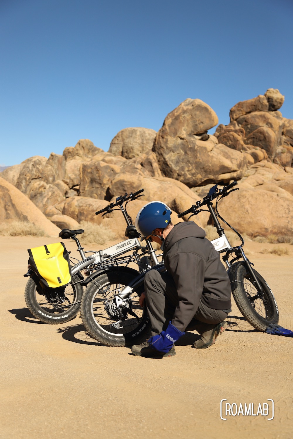 Man crouching next to bikes, looking at the tires, with golden boulders and a clear blue sky.