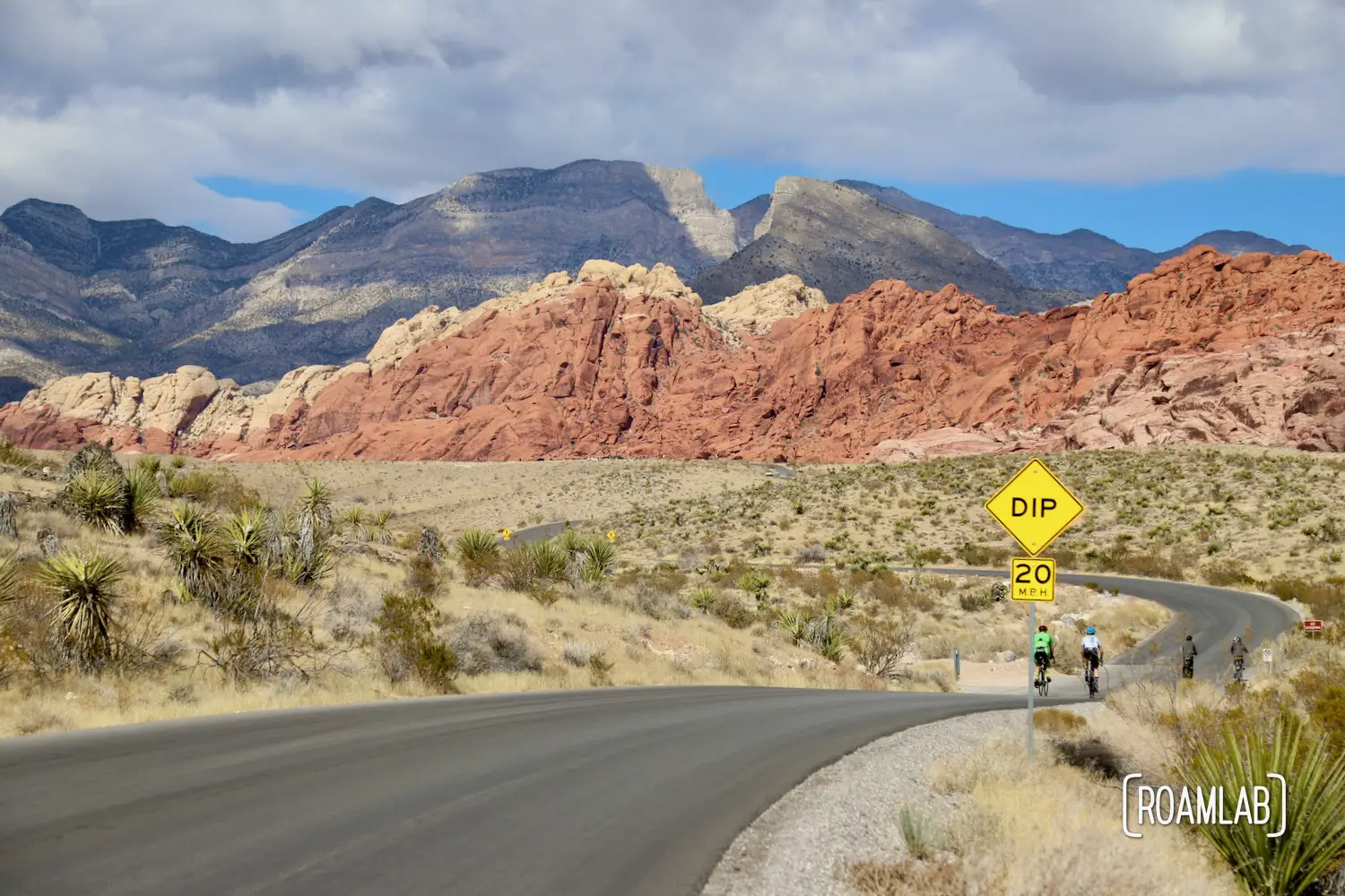 Two cyclists traveling down a paved road winding toward red rock cliffs.