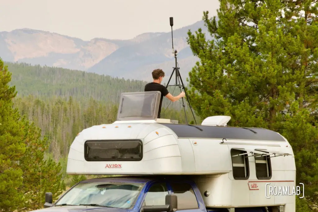 Man reaching through a roof hatch in an 1970 Avion C11 truck camper with a cell booster antenna set up on a tripod, surrounded with trees and mountains.