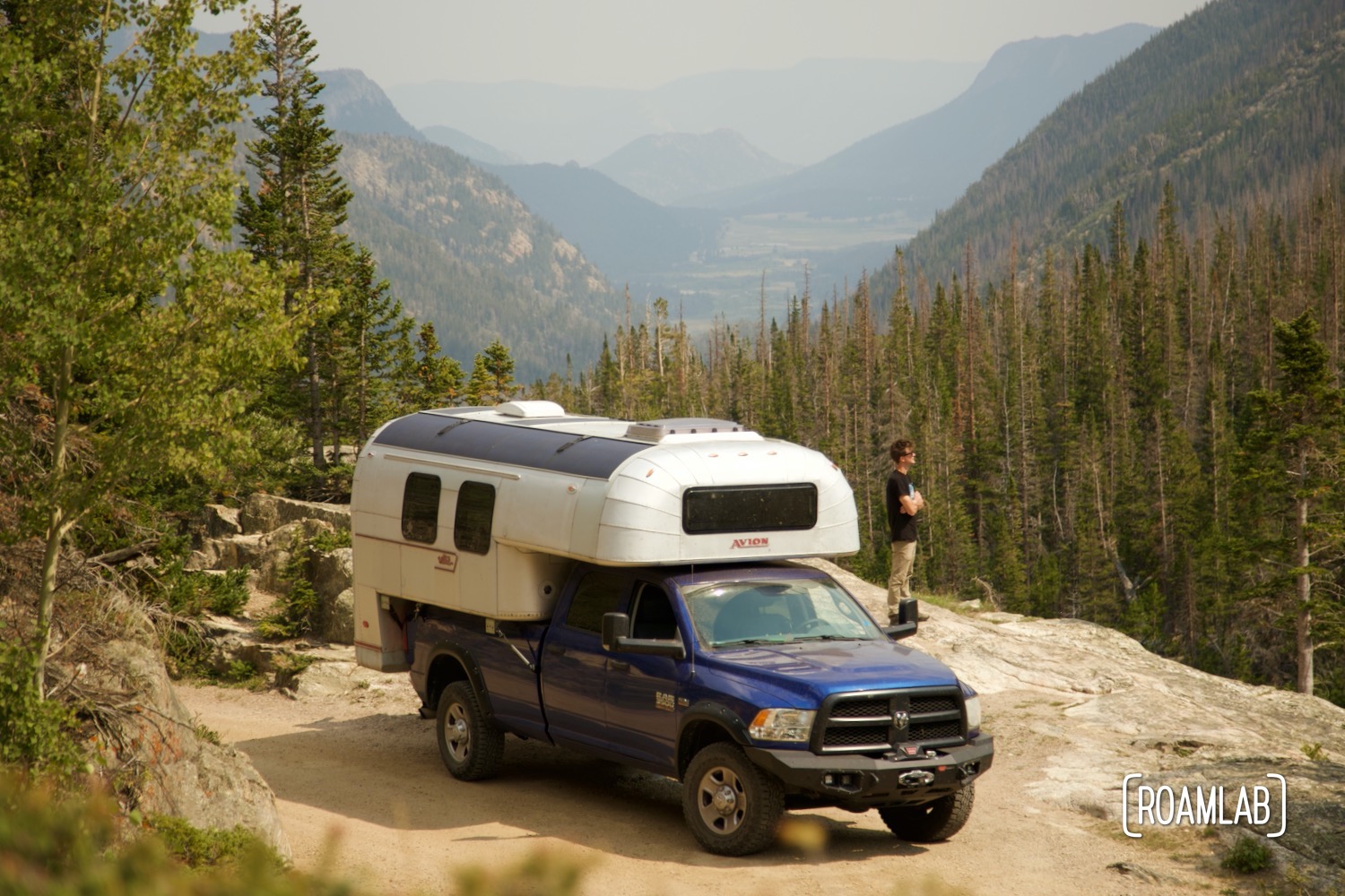 1970 Avion C11 truck camper parked along a dirt road with the driver looking out over a tree-lined mountain valley.