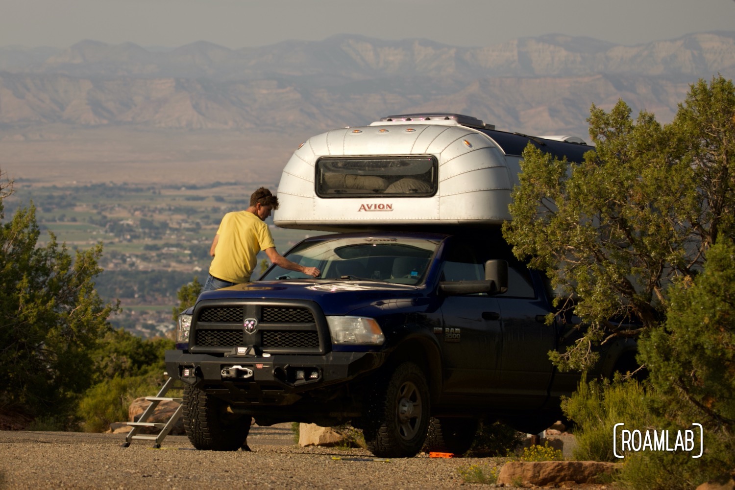 Man sitting on the hood of a blue truck with a 1970 Avion C11 truck camper at Saddlehorn Campground in Colorado National Monument.