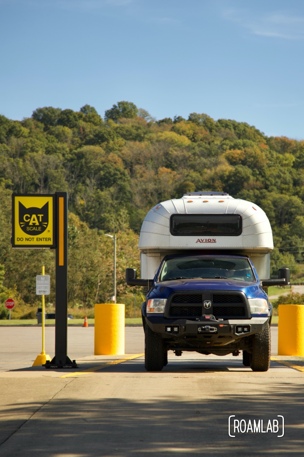 Vintage silver aluminum 1970 Avion C11 truck camper on a blue 2015 Ram 3500 truck parked on CAT scale pad with fall color forest hill in the background.