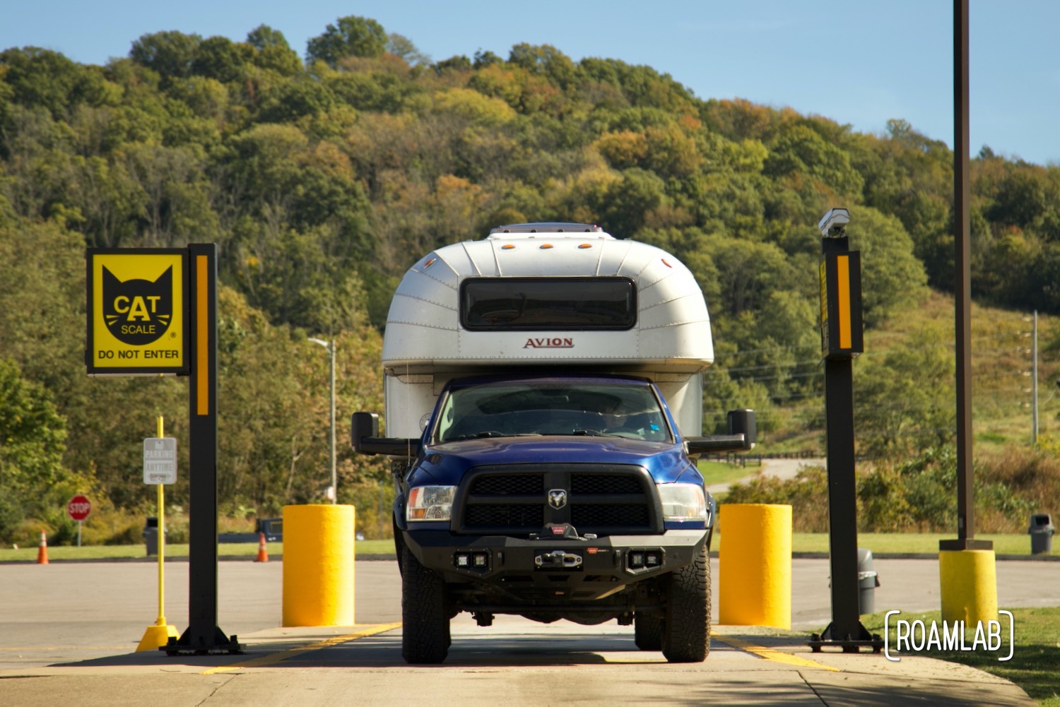 Vintage silver aluminum 1970 Avion C11 truck camper on a blue 2015 Ram 3500 truck parked on CAT scale pad with fall color forest hill in the background.