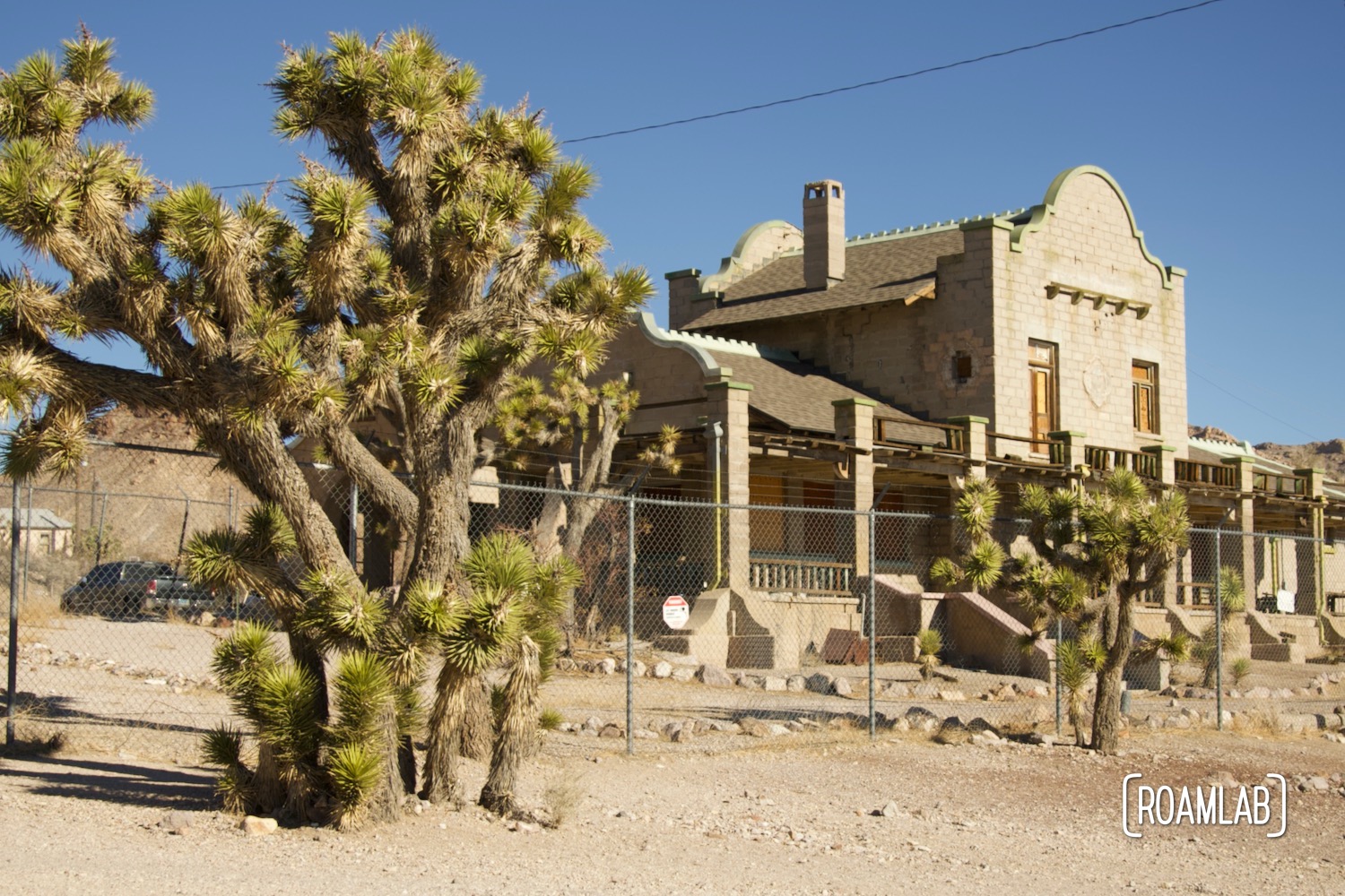 Pinkish stone Rhyolite train station surrounded by a chain link fence and bordered by joshua trees.