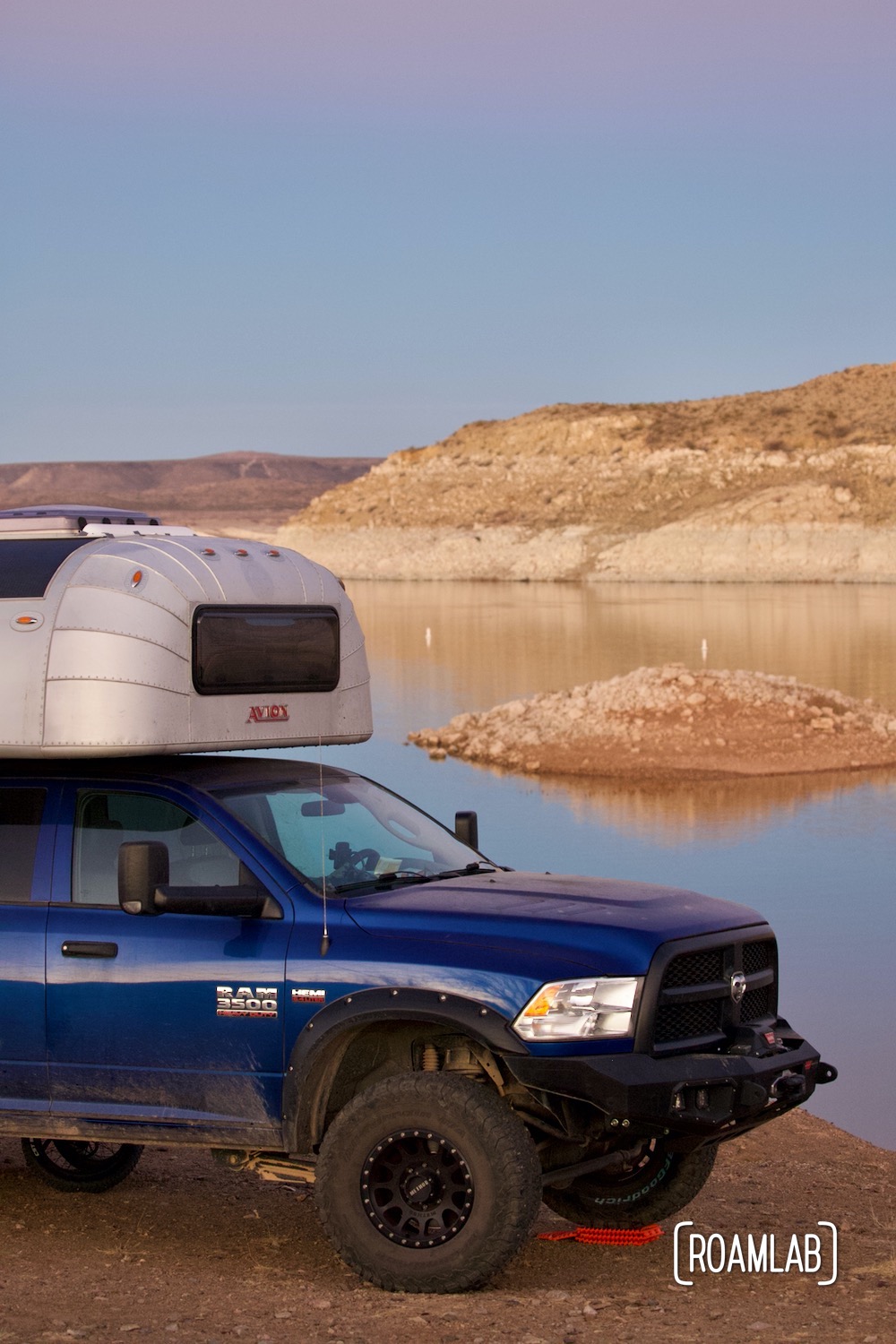 Front half of a 1970 Avion C11 truck camper and blue Ram truck parked in front of a couple lake bound islands at dusk.