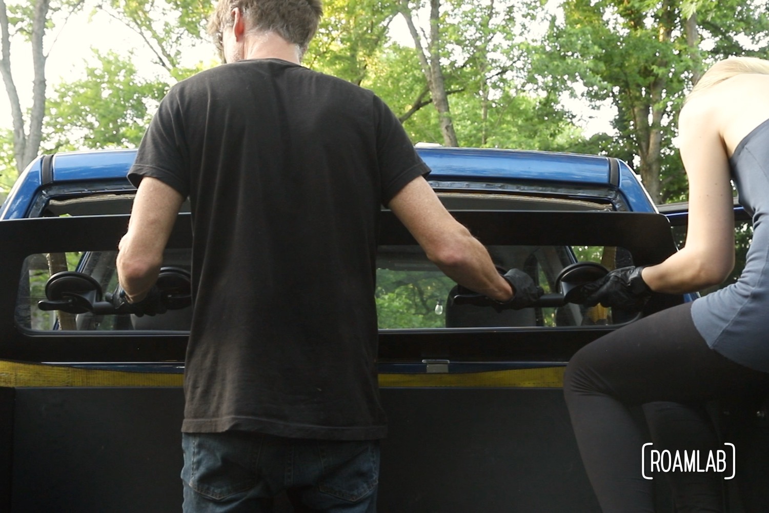 Man holding a truck rear windshield against the truck frame while a woman adjusts its position.