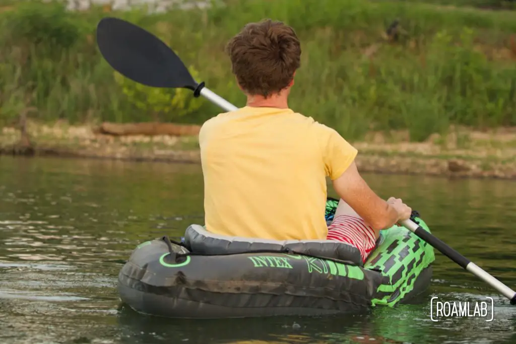 Man paddling in a kayak near a grassy shoreline.
