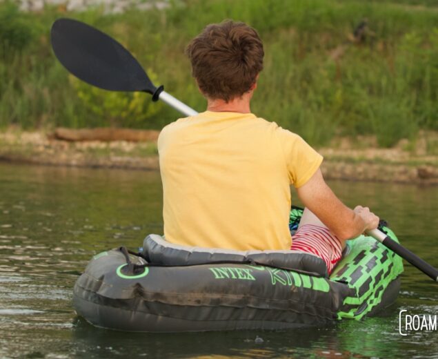 Man paddling in a kayak near a grassy shoreline.