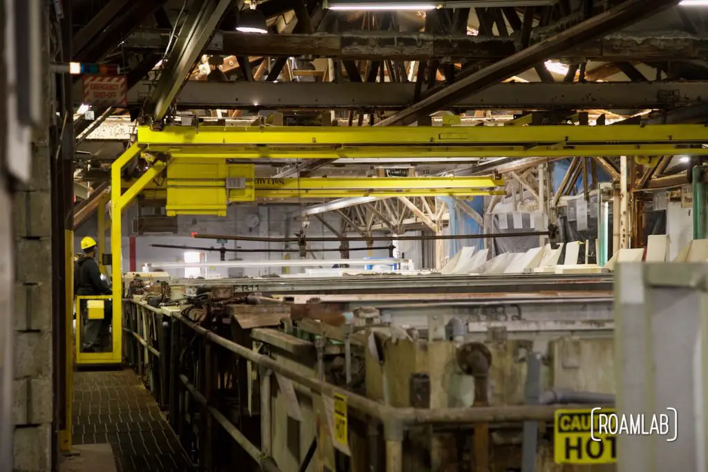 View inside a large warehouse filled by a row of vats and a yellow crane straddling the row.