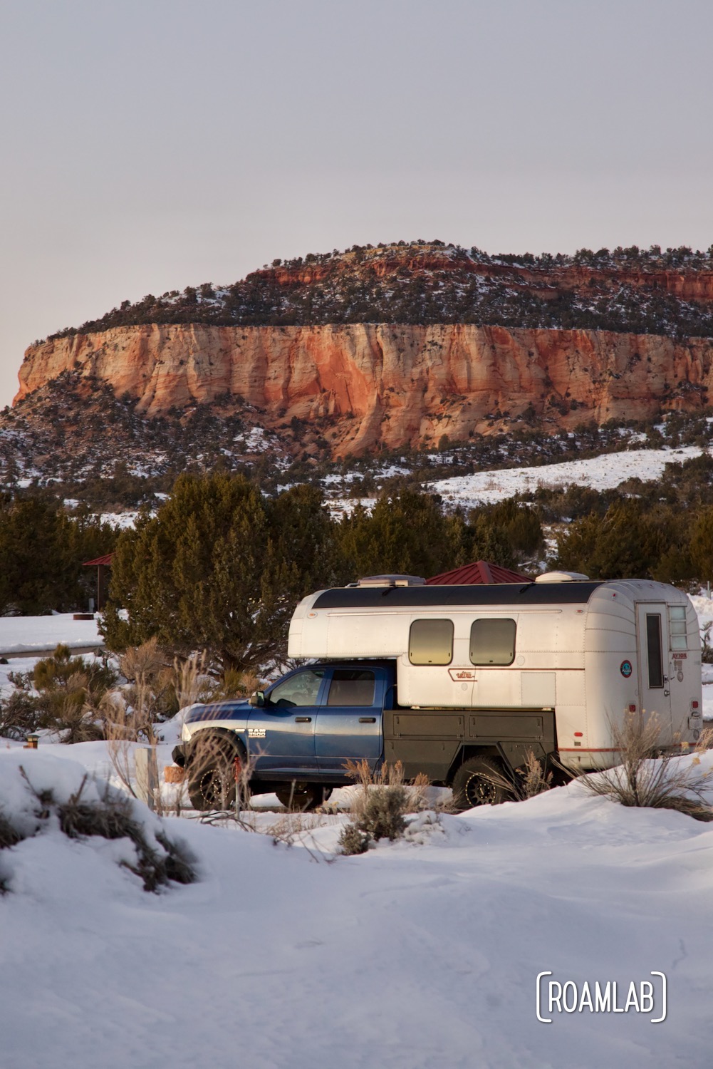 Avion C11 truck camper parked among snow drifts at sunset with pink cliffs rising in the distance at Coral Pink Sand Dunes State Park in Northern Arizona.
