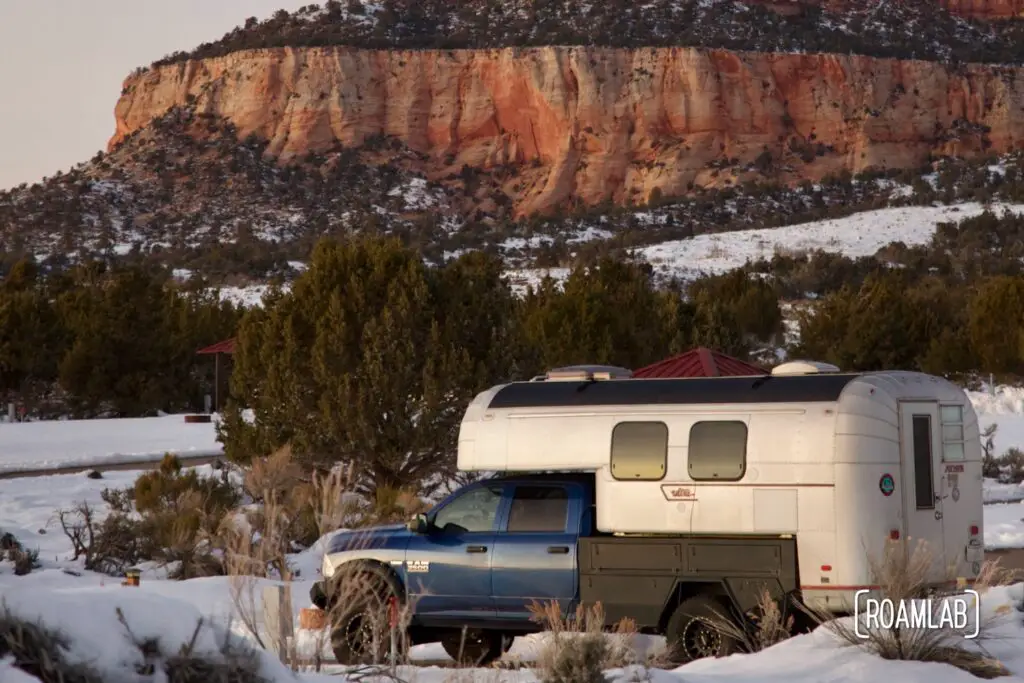 Avion C11 truck camper parked among snow drifts at sunset with pink cliffs rising in the distance at Coral Pink Sand Dunes State Park in Northern Arizona.