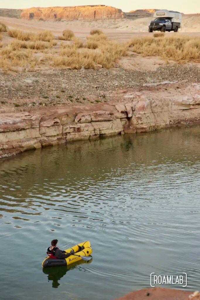 Man in a yellow raft paddling toward a pink butte with an Avion C11 truck camper parked at its edge.