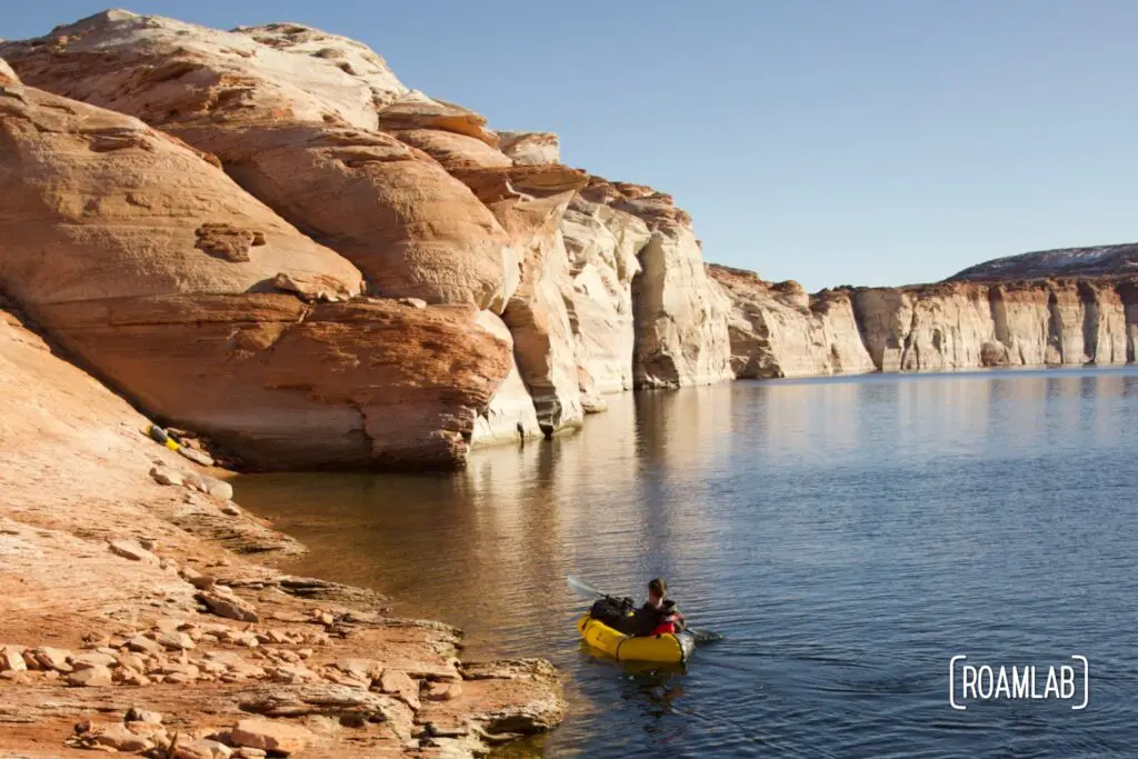 Man paddling in a yellow raft in the distance towards white cliffs along the south shore of Lake Powell in northern Arizona.