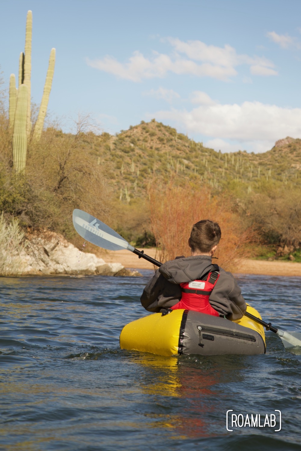 Man in a yellow raft paddling on a lake with desert brush and saguaro cactus in the background.