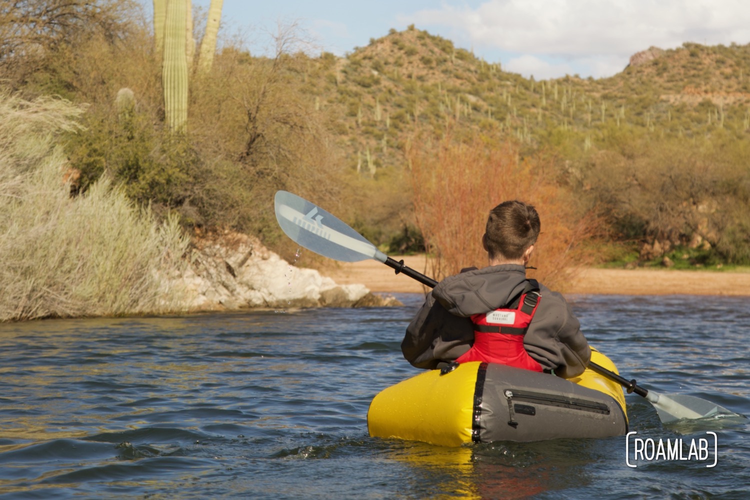 Man in a yellow raft paddling on a lake with desert brush and saguaro cactus in the background.