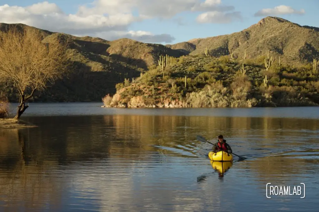 Man in a yellow raft with mountains in the background covered with desert brush and cactus.