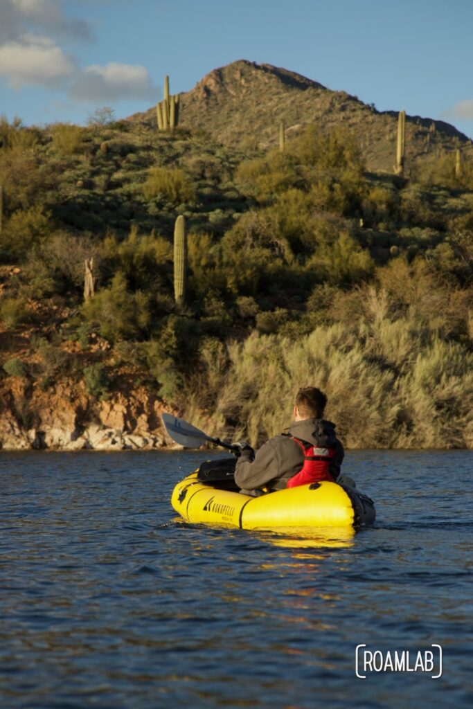 Man in a yellow raft paddling on a like with saguaro cactus in the background.