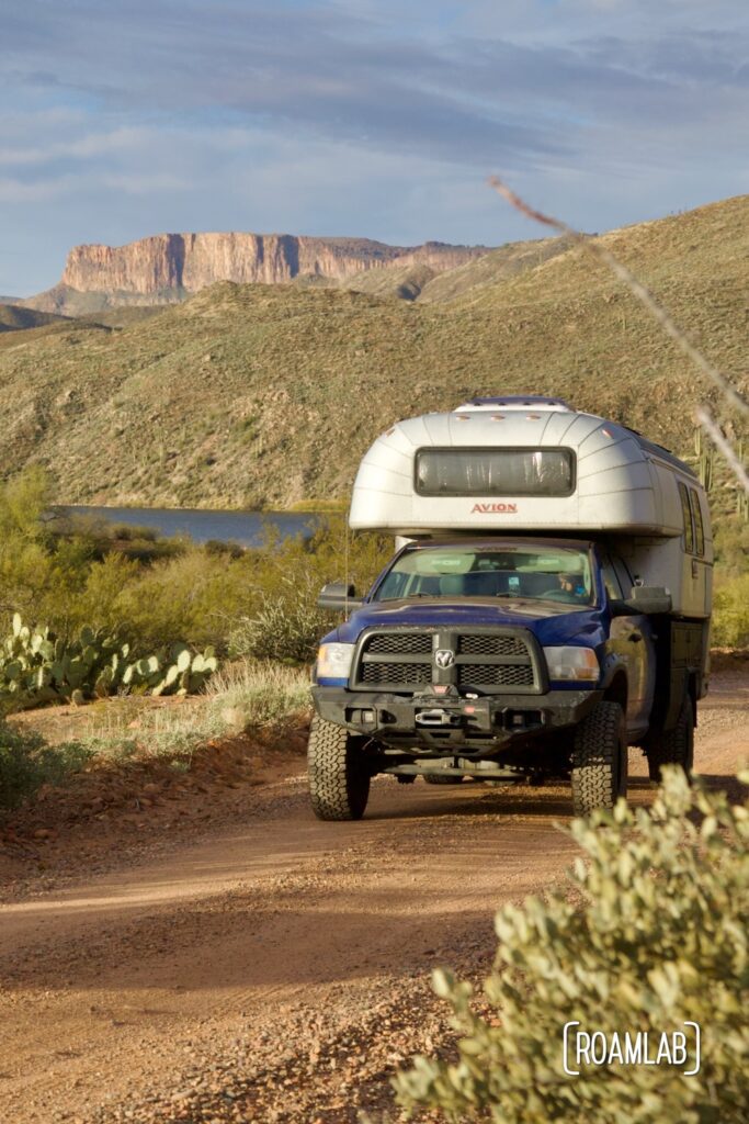 Avion C11 truck camper driving down a dirt road with a lake and cliffs in the background.