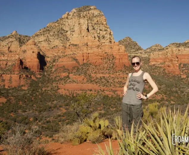Woman standing with hands on hip behind green agave with colorful red buttes in the background.