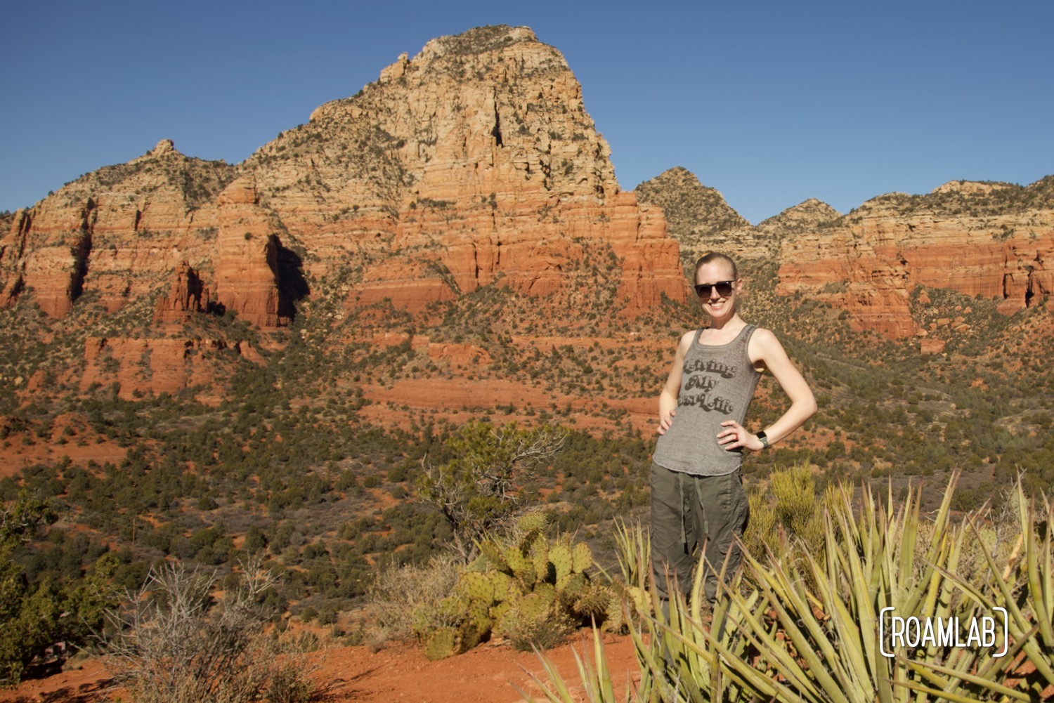 Woman standing with hands on hip behind green agave with colorful red buttes in the background.