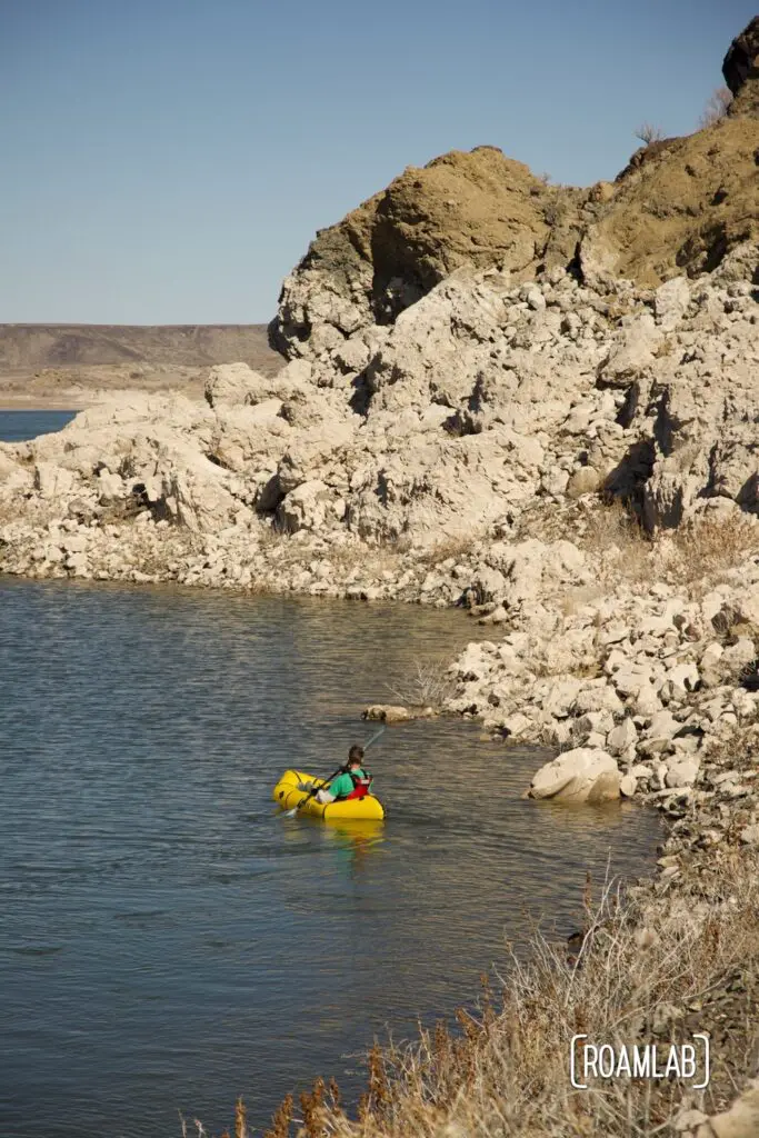 Man in a yellow Kokopelli packraft paddling along the rocky coastline of Elephant Butte Lake State Park.