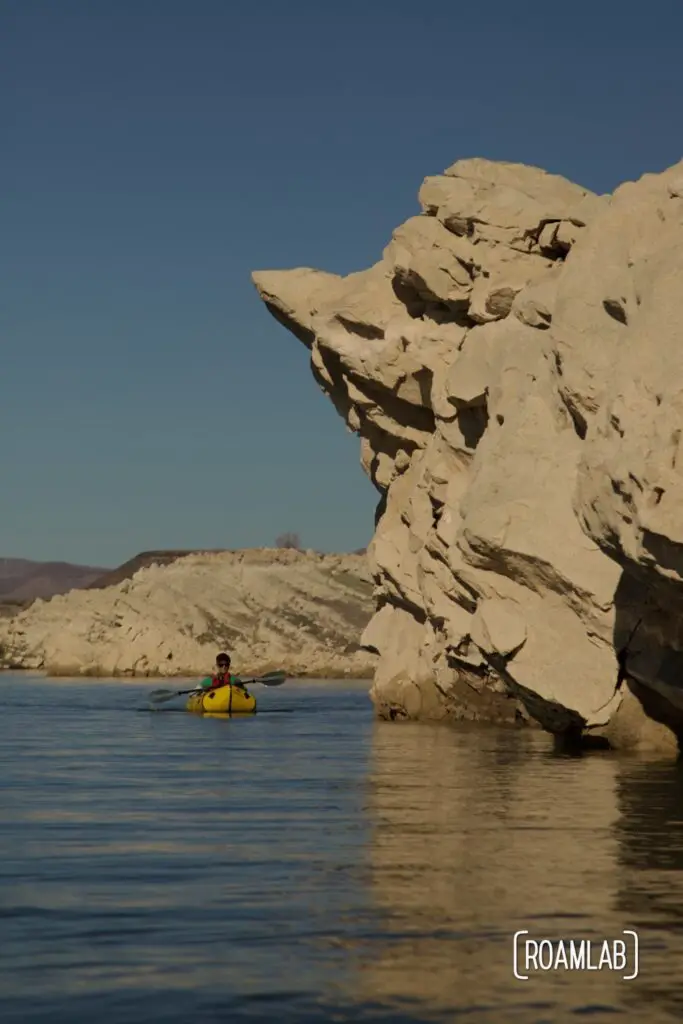 Man paddling a yellow packraft dwarfed by a golden cliff hanging over the water.