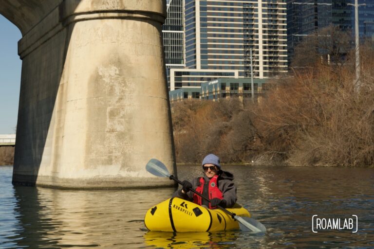 Man paddling by the pillar of Congress Avenue Bridge with Austin, Texas in the background.