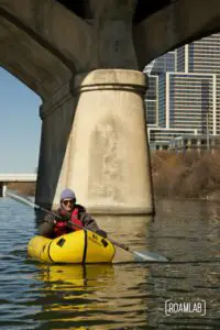 Man paddling by the pillar of Congress Avenue Bridge with Austin, Texas in the background.