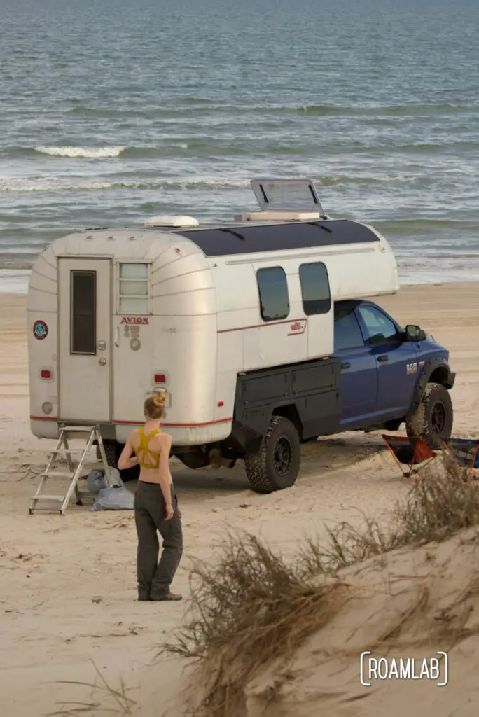 Woman walking around a 1970 Avion C11 truck camper parked on the beach of Padre Island National Seashore.