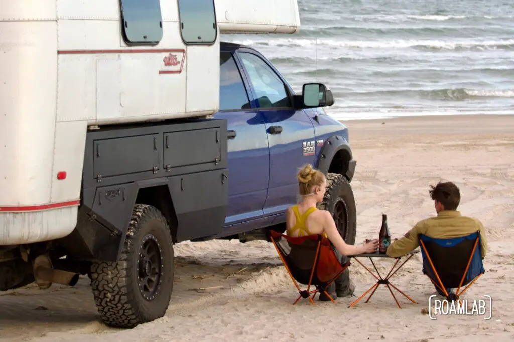 Man and woman sitting on the beach next to a 1970 Avion C11 truck camper on a blue Ram at Padre Island National Seashore.