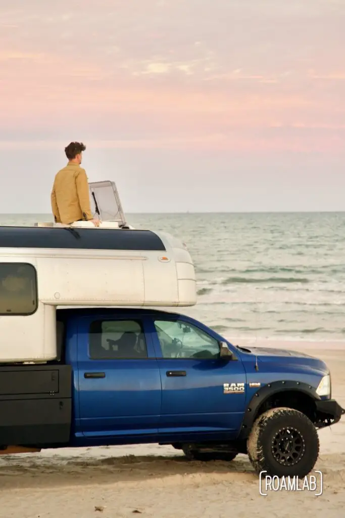 Man looking out a roof hatch of a 1970 Avion C11 truck camper at a sunset over the Gulf of Mexico while parked on the beach of Padre Island National Seashore in Texas.