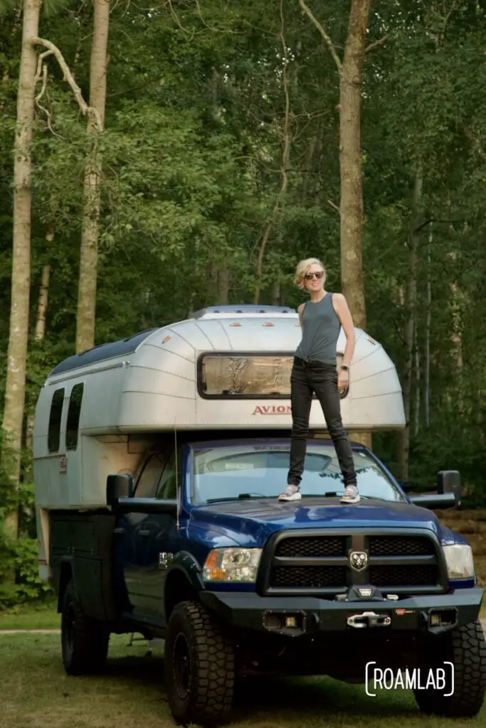 Woman standing on the hood of a 1970 Avion C11 truck camper parked in a Minnesota forest.