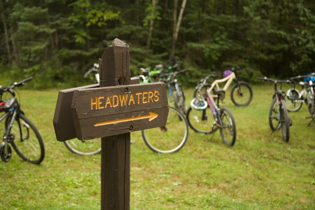 Wooden sign with "Headwaters" in yellow with an arrow and bicycles parked behind it.