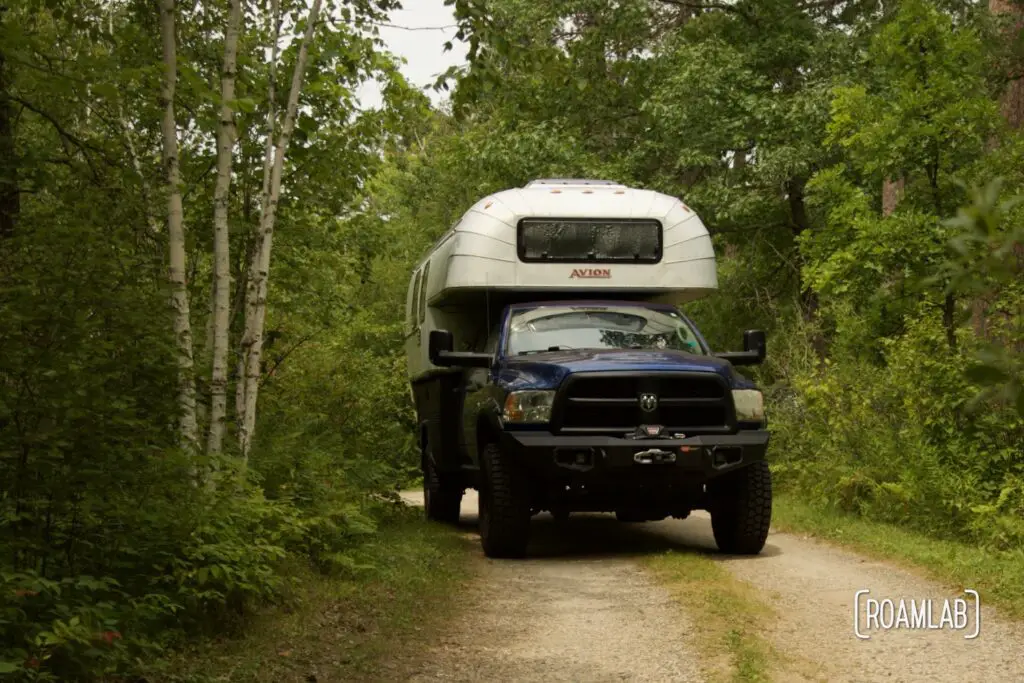 1970 Avion C11 truck camper driving down a gravel road in a lush forest.