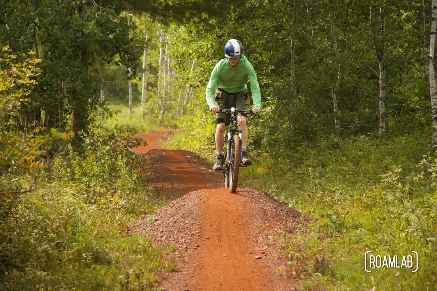 Man on a mountain bike peddling on red dirt single track in a forest.