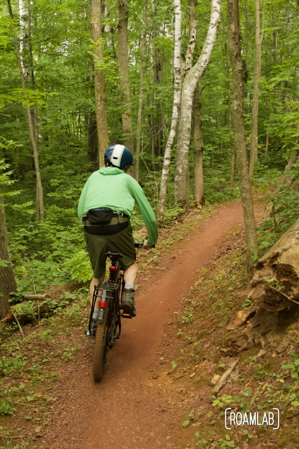 Man on a mountain bike peddling on red dirt single track in a forest.